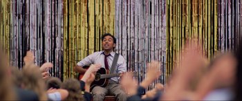Movie still from “Yesterday” (2019), directed by Danny Boyle – A man sitting in front of a group of people playing a guitar; Medium shot, Over the shoulder angle