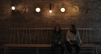 Movie still from “You Hurt My Feelings” (2023), directed by Nicole Holofcener – Two women sitting on a wooden bench in front of a brick wall; Wide shot, High angle