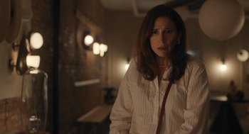 Movie still from “You Hurt My Feelings” (2023), directed by Nicole Holofcener – A woman standing in a dimly - lit room with her hands in her pockets; Medium shot, Over the shoulder angle