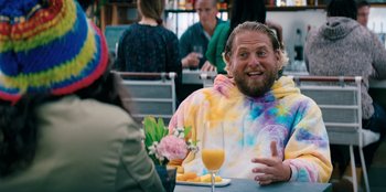 Movie still from “You People” (2023), directed by Kenya Barris – A man in a tie dye shirt sitting at a table with a glass of orange juice; Medium shot, Over the shoulder angle