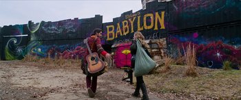 Movie still from “Zombieland: Double Tap” (2019), directed by Ruben Fleischer – A man holding a guitar in front of a woman in front of a building; Wide shot, Low angle