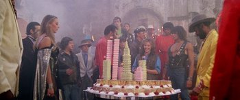Movie still from “1990: The Bronx Warriors” (1982), directed by Enzo G. Castellari – A group of people standing around a cake with towers on it; Medium shot, Over the shoulder angle