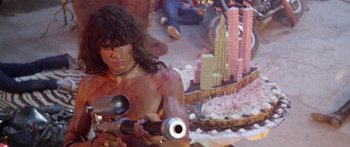 Movie still from “1990: The Bronx Warriors” (1982), directed by Enzo G. Castellari – A shirtless man holding a gun in front of a cake; Medium shot, Over the shoulder angle