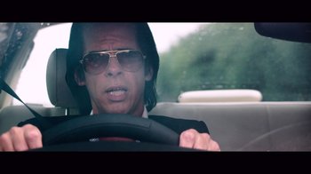 Movie still from “20,000 Days on Earth” (2014), directed by Iain Forsyth – A man driving a car wearing a suit and sunglasses; Close Up shot, Low angle