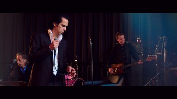 Movie still from “20,000 Days on Earth” (2014), directed by Iain Forsyth – A man holding a microphone while standing next to another man playing a guitar; Medium shot, Low angle