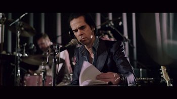 Movie still from “20,000 Days on Earth” (2014), directed by Iain Forsyth – A man sitting at a microphone with papers in front of him; Medium shot, Low angle