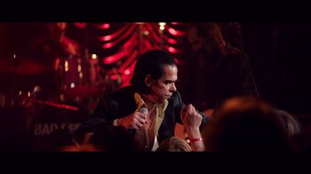 Movie still from “20,000 Days on Earth” (2014), directed by Iain Forsyth – A man holding a banana in his hand while sitting in front of a crowd; Medium shot, Over the shoulder angle