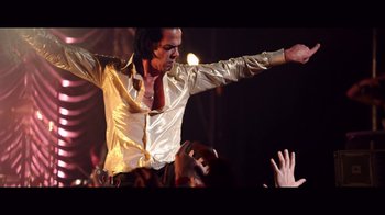 Movie still from “20,000 Days on Earth” (2014), directed by Iain Forsyth – A man in a gold shirt and red neck tie is on a stage; Medium shot, Low angle