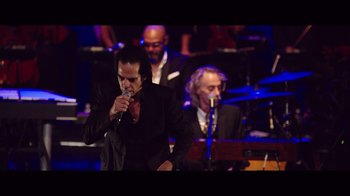 Movie still from “20,000 Days on Earth” (2014), directed by Iain Forsyth – A man singing into a microphone in front of an orchestra; Medium shot, Low angle