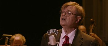 Movie still from “A Prairie Home Companion” (2006), directed by Robert Altman – An older man in a suit and red neck tie is speaking into a microphone; Close Up shot, Over the shoulder angle