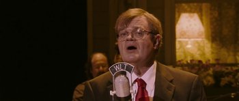 Movie still from “A Prairie Home Companion” (2006), directed by Robert Altman – An older man wearing a suit and tie talking into a microphone; Close Up shot, Low angle