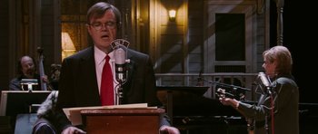 Movie still from “A Prairie Home Companion” (2006), directed by Robert Altman – A man in a suit and red tie standing in front of a microphone; Medium shot, Over the shoulder angle
