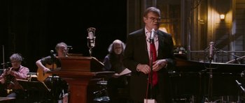 Movie still from “A Prairie Home Companion” (2006), directed by Robert Altman – A man wearing a suit and red tie standing at a microphone; Medium shot, Over the shoulder angle