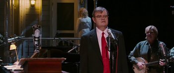 Movie still from “A Prairie Home Companion” (2006), directed by Robert Altman – A man wearing a suit and red tie standing at a microphone in front of a woman; Medium shot, Over the shoulder angle