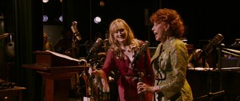 Movie still from “A Prairie Home Companion” (2006), directed by Robert Altman – Two women sing into microphones in front of a crowd; Medium shot, High angle