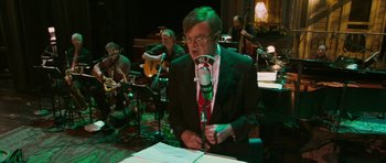 Movie still from “A Prairie Home Companion” (2006), directed by Robert Altman – A man holding a microphone in front of a group of people; Medium shot, High angle