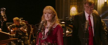 Movie still from “A Prairie Home Companion” (2006), directed by Robert Altman – A woman standing in front of a microphone in a room; Medium shot, Over the shoulder angle