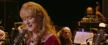 Movie still from “A Prairie Home Companion” (2006), directed by Robert Altman – A woman in a red dress is listening to music; Close Up shot, Over the shoulder angle