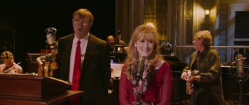 Movie still from “A Prairie Home Companion” (2006), directed by Robert Altman – A man and a woman singing into microphones in front of an orchestra; Medium shot, High angle