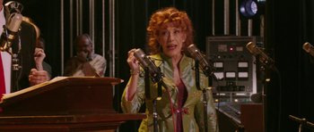 Movie still from “A Prairie Home Companion” (2006), directed by Robert Altman – An older woman with red curly hair talking into microphones; Medium shot, Over the shoulder angle