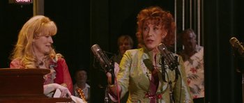 Movie still from “A Prairie Home Companion” (2006), directed by Robert Altman – An older woman is holding a microphone in front of a group of people; Medium shot, Over the shoulder angle