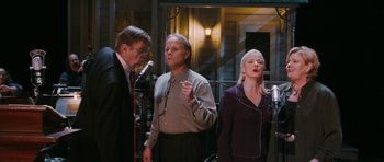 Movie still from “A Prairie Home Companion” (2006), directed by Robert Altman – A group of people singing into microphones in a room; Medium shot, High angle