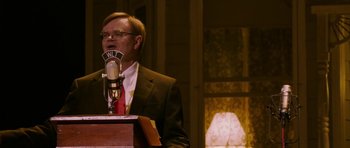 Movie still from “A Prairie Home Companion” (2006), directed by Robert Altman – A man in a suit and tie speaking into a microphone; Medium shot, Over the shoulder angle