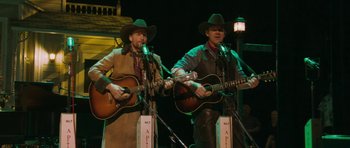 Movie still from “A Prairie Home Companion” (2006), directed by Robert Altman – A couple of men in cowboy hats playing guitars on a stage; Medium shot, Over the shoulder angle