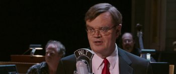 Movie still from “A Prairie Home Companion” (2006), directed by Robert Altman – An older man in a suit and red neck tie is talking into a microphone; Close Up shot, Over the shoulder angle