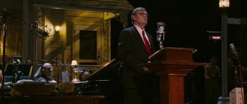 Movie still from “A Prairie Home Companion” (2006), directed by Robert Altman – A man standing at a podium in front of a microphone; Medium shot, Low angle