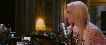 Movie still from “A Prairie Home Companion” (2006), directed by Robert Altman – A woman with blonde hair standing in front of a microphone; Medium shot, Over the shoulder angle