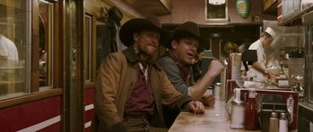 Movie still from “A Prairie Home Companion” (2006), directed by Robert Altman – Two men sitting at a table in cowboy hats; Medium shot, Over the shoulder angle