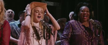 Movie still from “A Prairie Home Companion” (2006), directed by Robert Altman – A woman singing into a microphone in front of a group of people; Medium shot, Over the shoulder angle