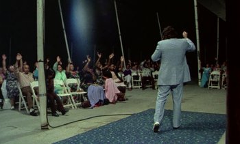 Movie still from “Alambrista!” (1977), directed by Robert M. Young – A crowd of people sitting and standing in front of a man in a blue suit; Wide shot, High angle