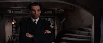 Movie still from “Anastasia” (1956), directed by Anatole Litvak – A man in a suit and tie with his arms crossed in front of a staircase; Medium shot, Low angle