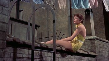 Movie still from “Artists and Models” (1955), directed by Frank Tashlin – A woman sitting on a brick wall in front of a building; Medium shot, Low angle