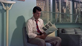 Movie still from “Artists and Models” (1955), directed by Frank Tashlin – A man sitting on a chair while reading a magazine; Medium shot, Over the shoulder angle