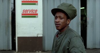 Movie still from “Babylon” (1980), directed by Franco Rosso – A man wearing a green hat standing in front of a gas station; Close Up shot, Low angle