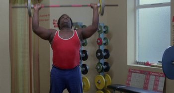 Movie still from “Babylon” (1980), directed by Franco Rosso – A man in a red tank top is lifting a barbell; Medium shot, Low angle