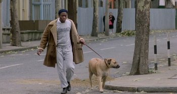 Movie still from “Babylon” (1980), directed by Franco Rosso – A man walking a dog on a leash down a street; Wide shot, High angle