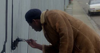 Movie still from “Babylon” (1980), directed by Franco Rosso – A man in a brown jacket looking at a cell phone; Medium shot, Over the shoulder angle