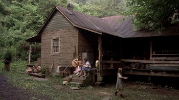 Movie still from “Blaze” (1989), directed by Ron Shelton – A woman sitting on a porch playing a guitar while two children watch; Wide shot, High angle