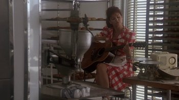 Movie still from “Blaze” (1989), directed by Ron Shelton – A woman in a red and white checkered dress playing an acoustic guitar; Wide shot, Over the shoulder angle