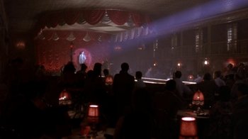 Movie still from “Blaze” (1989), directed by Ron Shelton – People are sitting at a table in front of a stage; Wide shot, High angle