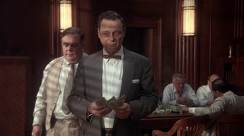 Movie still from “Blaze” (1989), directed by Ron Shelton – A man in a suit and bow tie holding money in front of other men in suits; Medium shot, Low angle