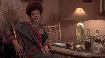 Movie still from “Blaze” (1989), directed by Ron Shelton – A woman sitting at a table in a room with a lamp; Medium shot, Over the shoulder angle