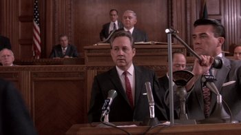 Movie still from “Blaze” (1989), directed by Ron Shelton – A man in a suit and tie sitting at a podium in front of microphones in a courtroom; Medium shot, Low angle