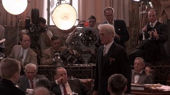 Movie still from “Blaze” (1989), directed by Ron Shelton – A man in a suit and tie standing in front of a camera; Medium shot, Over the shoulder angle