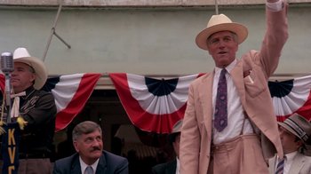 Movie still from “Blaze” (1989), directed by Ron Shelton – A man wearing a suit and a cowboy hat stands in front of an american flag; Medium shot, Low angle