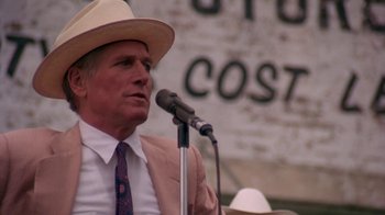 Movie still from “Blaze” (1989), directed by Ron Shelton – A man wearing a cowboy hat is speaking into a microphone; Medium shot, Over the shoulder angle